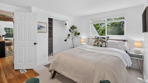 A bedroom with a large bed, a window with a view of greenery, a bedside table with a lamp, and a door leading to another room.