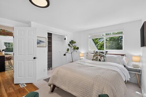 A bedroom with a large bed, a window with a view of greenery, a bedside table with a lamp, and a door leading to another room.