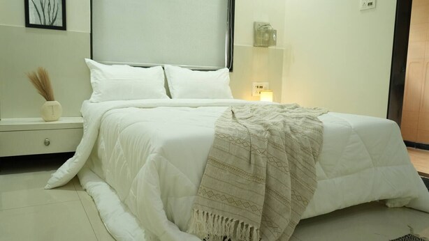 A neatly made bed with a white duvet and a beige throw blanket, a bedside table with a vase, and a wall-mounted shelf with a small item.
