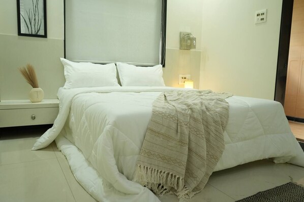 A neatly made bed with a white duvet and a beige throw blanket, a bedside table with a vase, and a wall-mounted shelf with a small item.