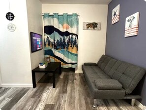 A living room with a grey sofa, a small table, a TV, and a curtain with a nature scene. - Apto Familiar cerca de Universal y Disney (Altamonte Springs)