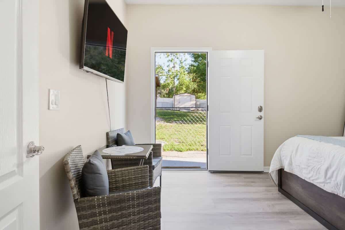 A bedroom with a bed, a television mounted on the wall, a wicker chair, and a door leading to an outdoor area.