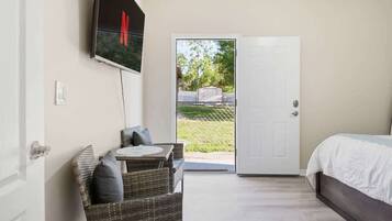A bedroom with a bed, a television mounted on the wall, a wicker chair, and a door leading to an outdoor area.