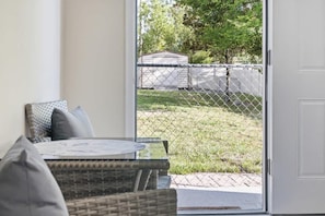 Una habitación con un sofá de mimbre y una mesa de vidrio, una puerta corrediza que da a un área exterior cercada y una puerta blanca.