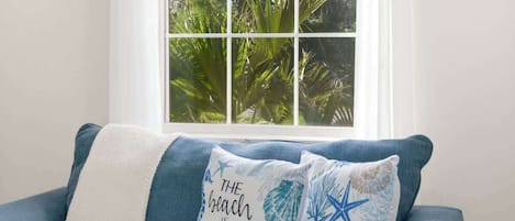 A blue sofa with two decorative pillows, one with a beach-themed design, positioned in front of a window with a view of green foliage.