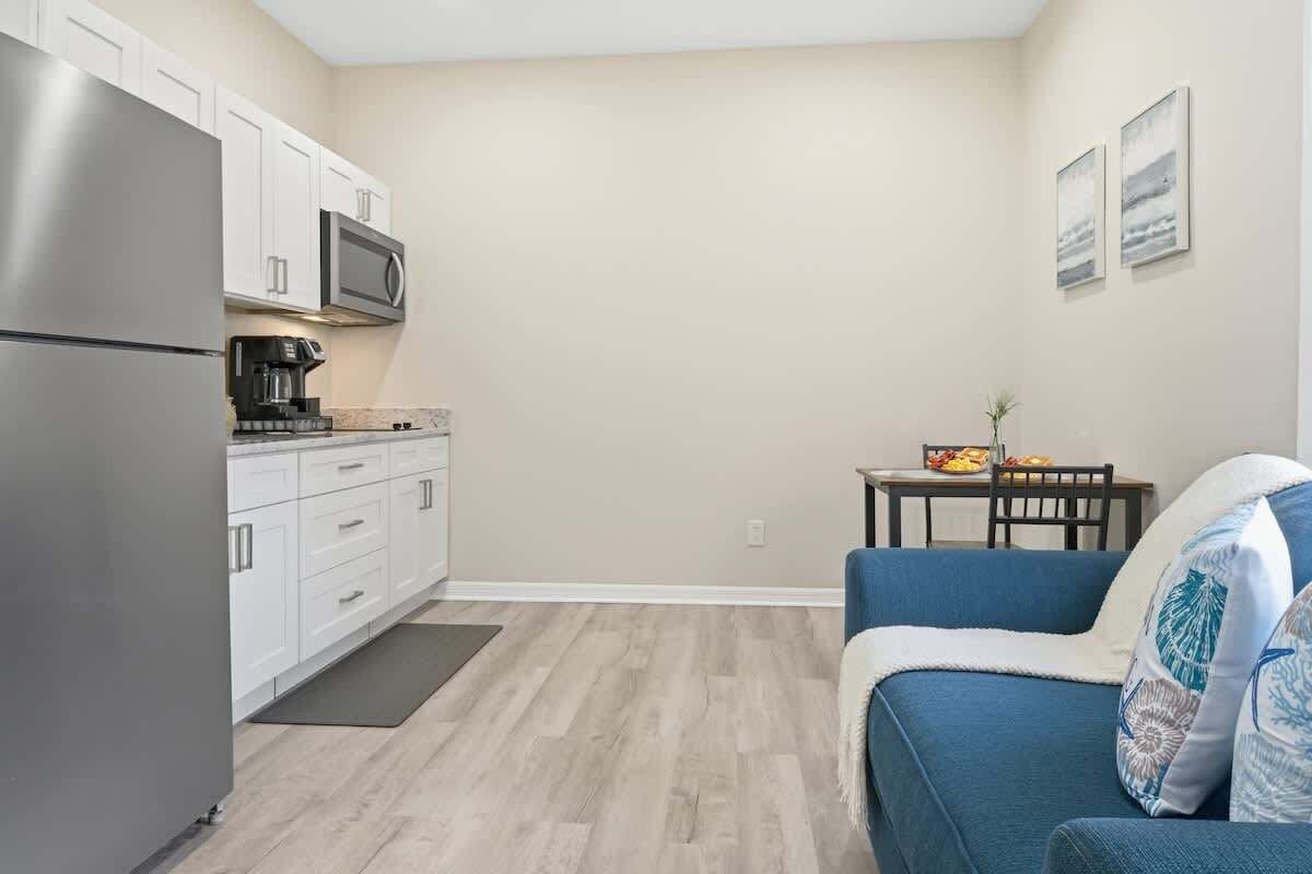 A compact kitchen with white cabinets, a stainless steel refrigerator, and a microwave. There's a dining table with chairs and a blue sofa with patterned cushions.