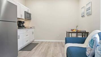 A compact kitchen with white cabinets, a stainless steel refrigerator, and a microwave. There's a dining table with chairs and a blue sofa with patterned cushions.