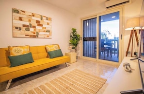 A yellow sofa with patterned cushions, a woven rug, a potted plant, and a sliding glass door leading to an outdoor area. - Costa Azul Apt: Pool Access Near La Vela Mall (Costa Azul)