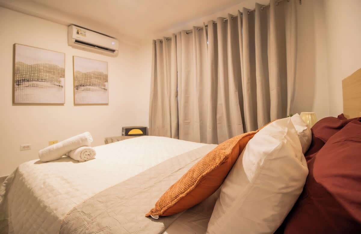 Una camera d'albergo con un letto, un cuscino, un asciugamano arrotolato, un condizionatore d'aria e delle tende.