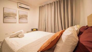 Una camera d'albergo con un letto, un cuscino, un asciugamano arrotolato, un condizionatore d'aria e delle tende.