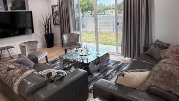 A modern living room with a sectional sofa, a glass coffee table, and a flat-screen TV mounted on the wall.