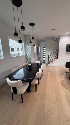 Ein moderner Essbereich mit einem langen Tisch, mehreren Stühlen, Pendelleuchten und einer Treppe im Hintergrund.