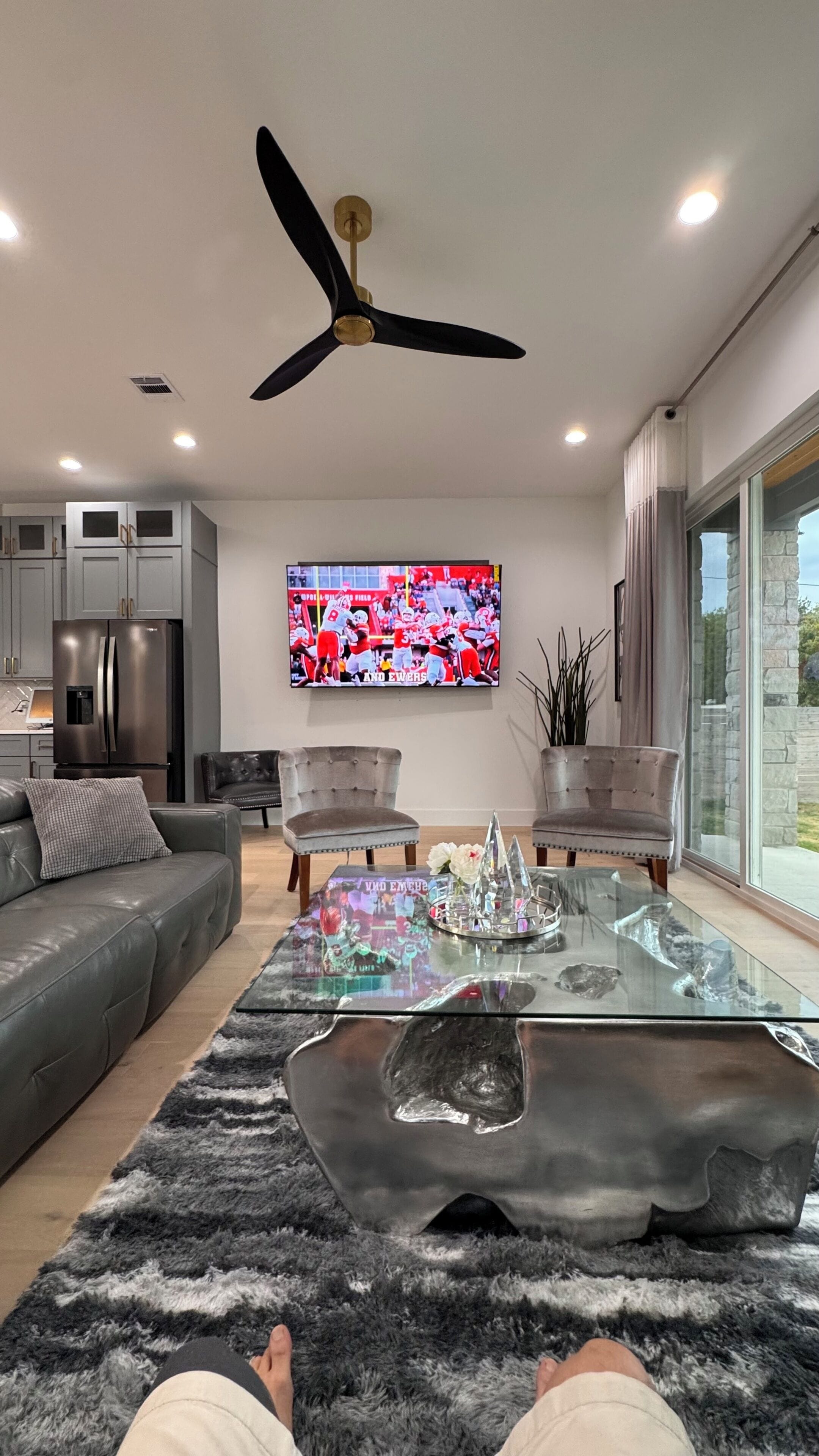 A modern living room with a flat-screen TV, a ceiling fan, and a glass coffee table.