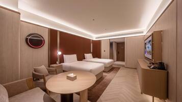 A modern hotel room with a round table, two beds, a sofa, and a TV on a stand.