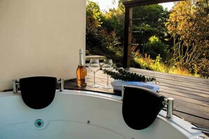 A hot tub with two black seats, a bottle of wine, and two glasses on a wooden deck with a view of a garden.