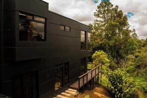A modern two-story building with a dark exterior, large windows, and a wooden deck leading to a lush green landscape.
