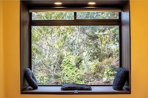 Ein Fenster mit Blick auf dichtes Grün, ein Kissen auf der Fensterbank und ein dunkler Rahmen.