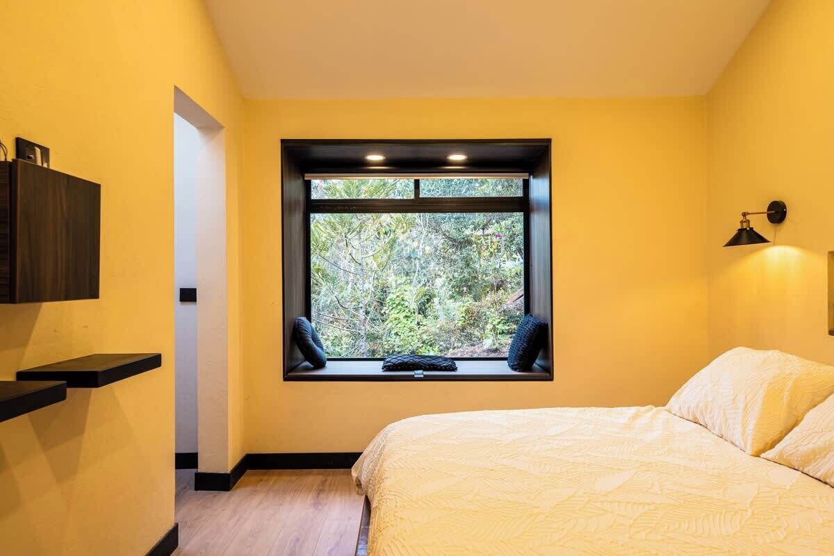 Un dormitorio con cama, ventana con vista a follaje, estante con objetos y una lámpara de pared.
