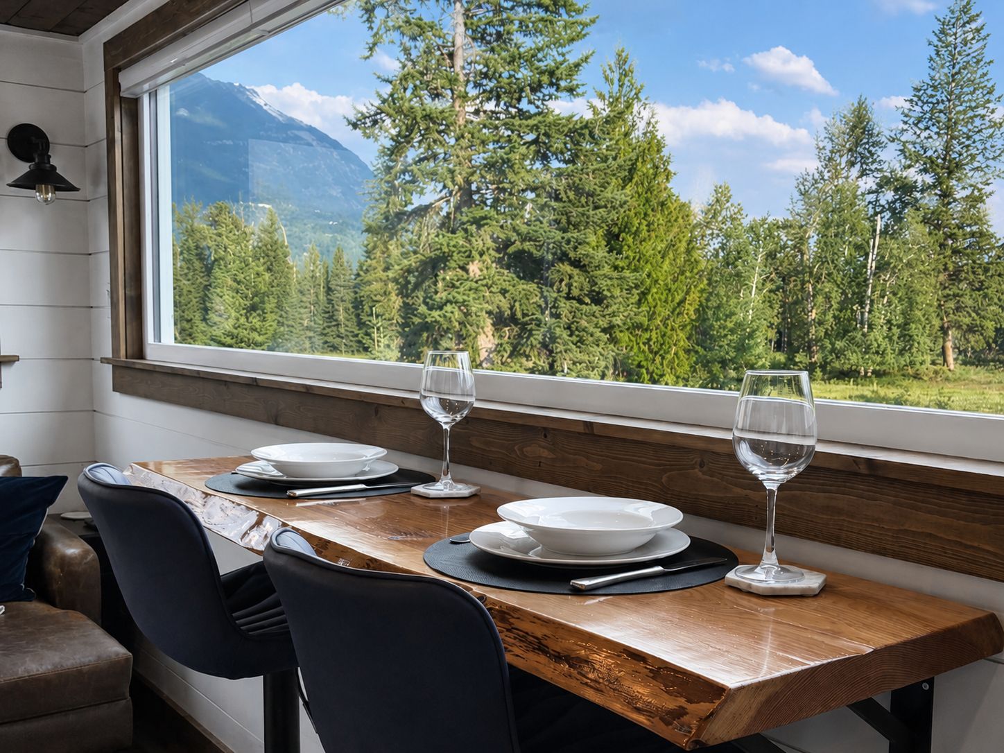 Une table à manger, avec assiettes et verres, placée devant une fenêtre offrant une vue sur les montagnes.