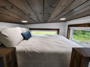 Un dormitorio con techo de madera, cama con almohada color beige y azul, y ventana con vista a follaje.