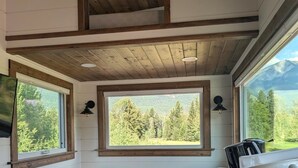 A cabin interior with a wooden ceiling, two windows with mountain views, and a table with a chair. - Nicholson Tiny Home with Hot Tub & Mountain Views (Golden)