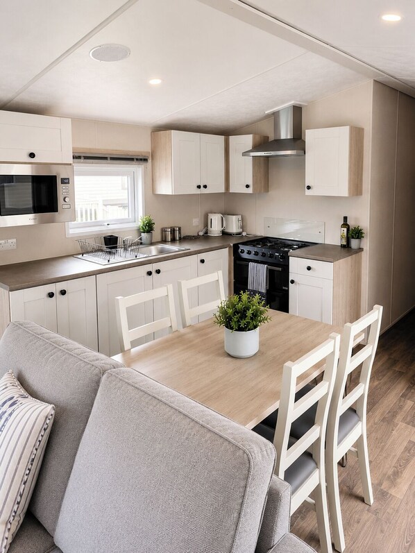 A modern kitchen with white cabinets, a microwave, a stove, and a dining area with a table and chairs. - 3 Bedroom Cabin Sleeps 6 - Shared Pool & Hot Tub (Malton)