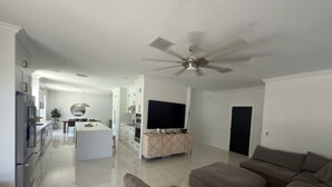 Ein modernes Wohnzimmer mit einem großen grauen Ecksofa, einer Küchenzeile mit weißen Schränken und einem Deckenventilator.