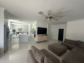 A modern living room with a large grey sectional sofa, a kitchen area with white cabinetry, and a ceiling fan. - Luxurious modern estate minutes from Disney (Davenport)