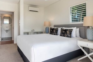 A modern bedroom with a large bed, bedside lamps, a window with blinds, and an air conditioning unit.