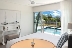 Habitación de hotel con cama, escritorio, silla, ventilador de techo y ventana grande con vista a una piscina y vegetación.