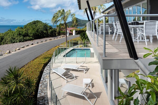 A poolside area with lounge chairs, a glass-enclosed pool, and a view of the ocean and hills.
