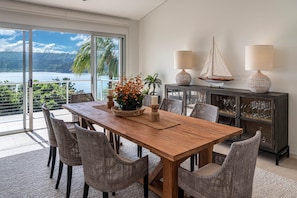 Un comedor con mesa de madera, sillas de mimbre, vista al océano y un aparador con objetos decorativos.