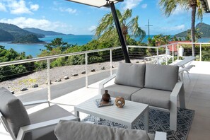A balcony with a sofa, a small table, and a view of the sea and mountains.