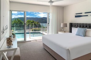 A hotel room with a large bed, a desk, a glass coffee table, and a view of a swimming pool and mountains.