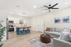A modern living room with a grey sofa, a coffee table, and a dining area with bar stools. - Fall Deals + Snowbird Rates | Near Downtown & Historic St. Andrews Bay (Panama City)