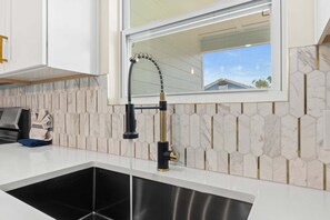 A modern kitchen with a stainless steel sink, a black faucet, and a window with a view of a house. - Fall Deals + Snowbird Rates | Near Downtown & Historic St. Andrews Bay (Panama City)