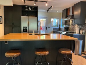 A modern kitchen with a central island, bar stools, a refrigerator, and a 'EAT' sign above the door. - Tree house steps from the river (Bend)