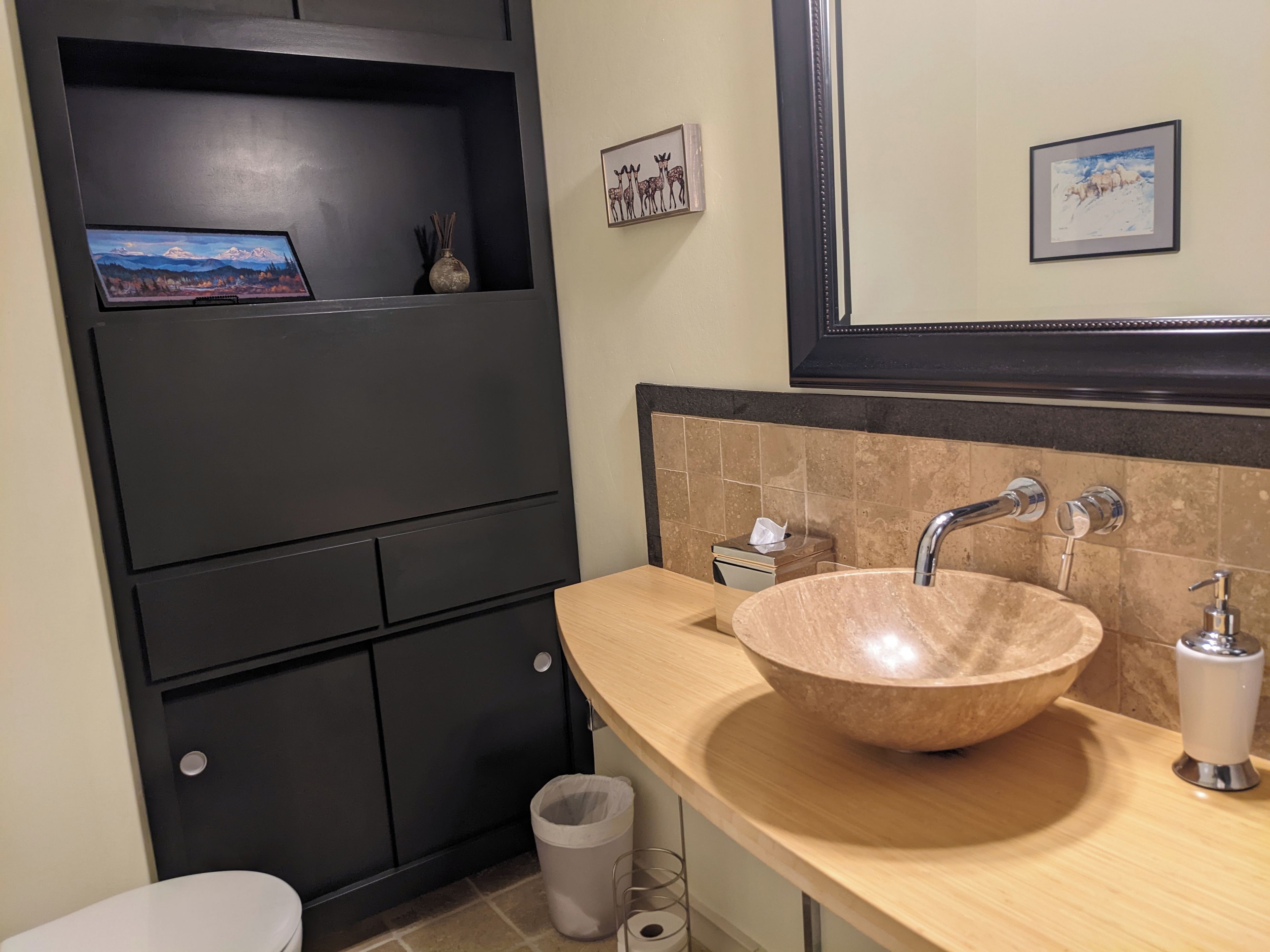Et moderne badeværelse med en stor beige vask, et spejl og et toilet. Der er pynteting på væggen og en skraldespand.