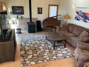 A living room with a stone-patterned rug, a brown sofa, a piano, a fireplace, and a television. - Tree house steps from the river (Bend)