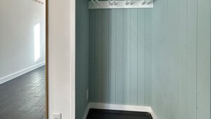 A corner of a room with a light blue wall, a white coat rack with hooks, a mirror with a gold frame, and a dark tiled floor. - Zeni House (Otaru City)