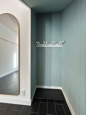 A corner of a room with a light blue wall, a white coat rack with hooks, a mirror with a gold frame, and a dark tiled floor.