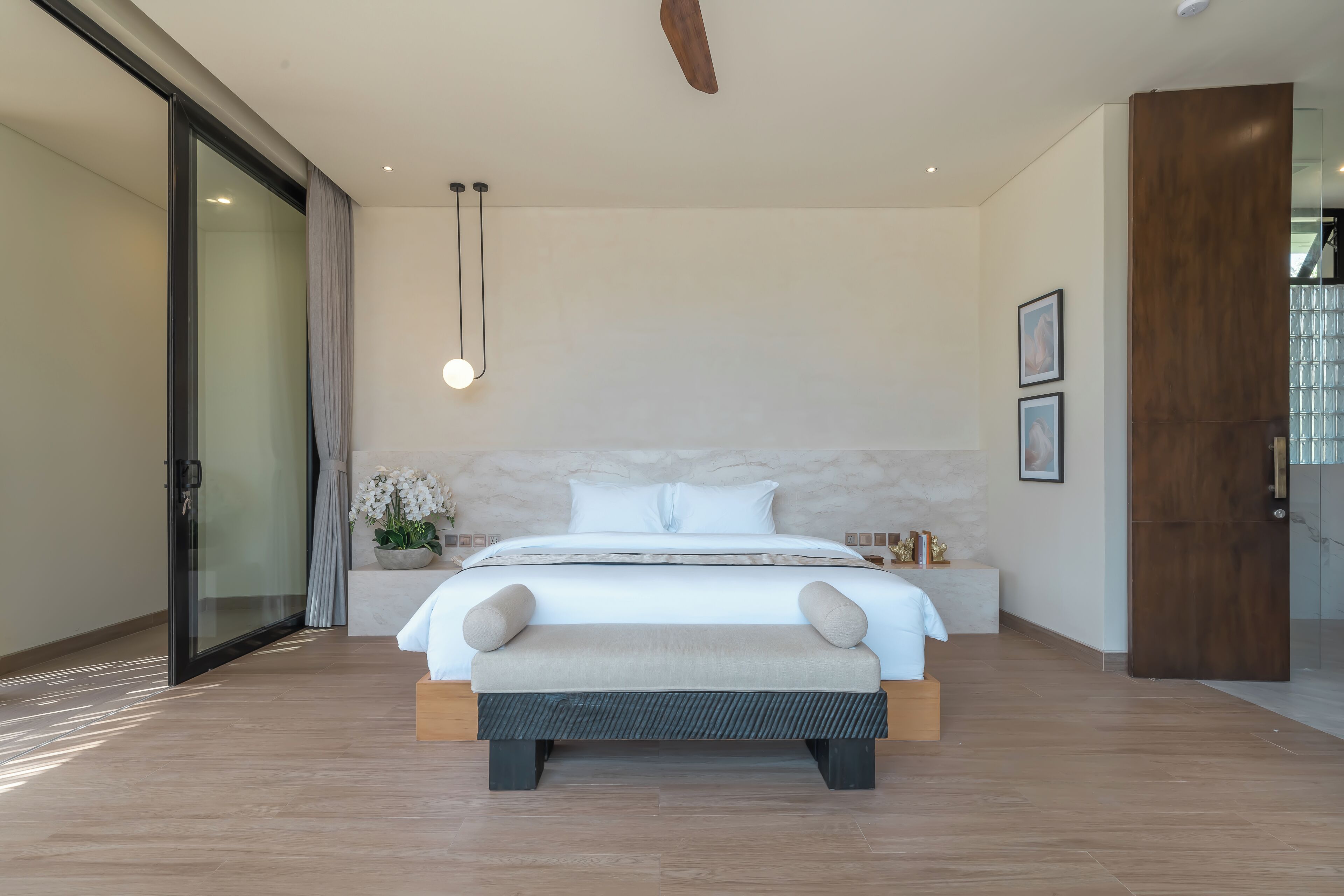 A modern bedroom with a large bed, a bedside table, a nightstand, a wall-mounted light, and a large window.