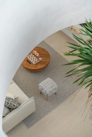 Ein Zimmer mit einem runden Holztisch, einem Schachbrettspiel, einer Fernbedienung, einem gewebten Teppich, einem grauen Pouf und einer Topfpflanze.