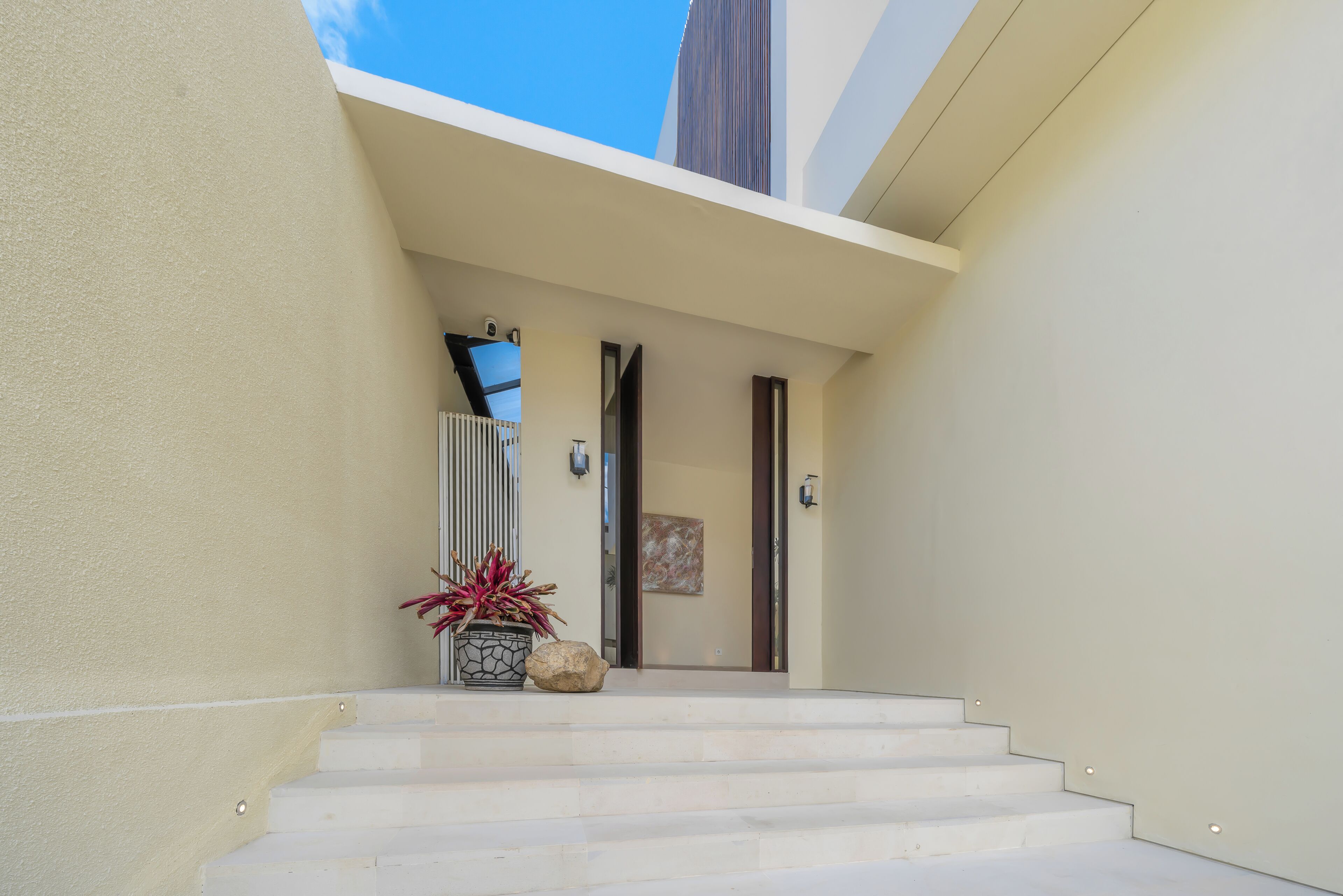 A modern building entrance with a set of stairs leading to a door, a potted plant, and a security camera.