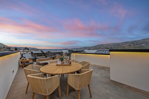 A rooftop terrace with a round wooden table and wicker chairs, overlooking a cityscape during sunset.