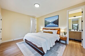 A bedroom with a large bed, bedside lamps, a nightstand, a painting of a lake, and a view into the bathroom.