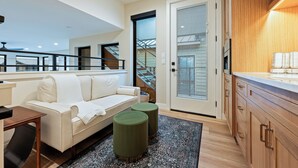 A modern living room with a white sofa, green ottomans, and a kitchen area with wooden cabinets and a countertop.