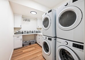 A laundry room with stacked front-loading washing machines, a drying rack, and storage bins.