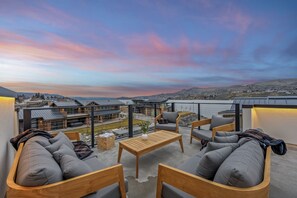 A rooftop terrace with a wooden table and grey sofas, overlooking a cityscape and hills.