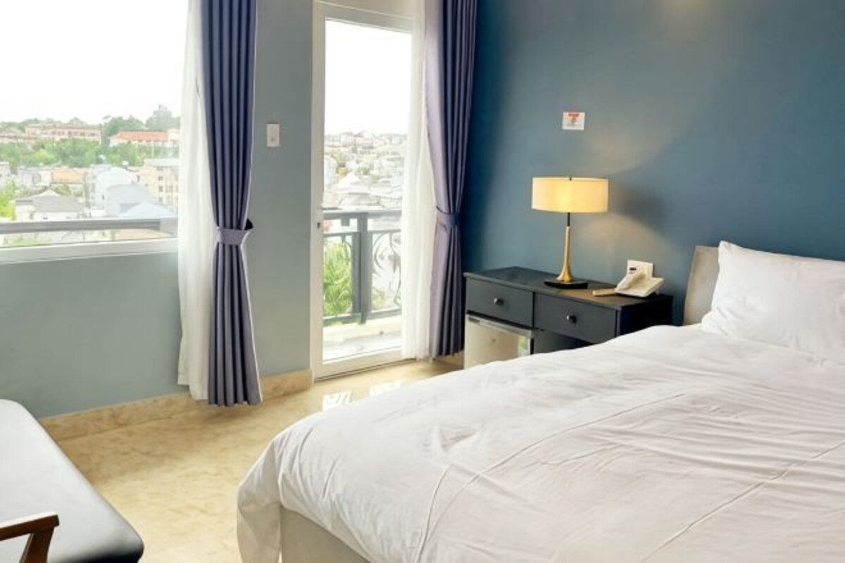 Un cuarto de hotel con cama, mesita de noche con lámpara, teléfono y vista de la ciudad a través de una ventana con cortinas.